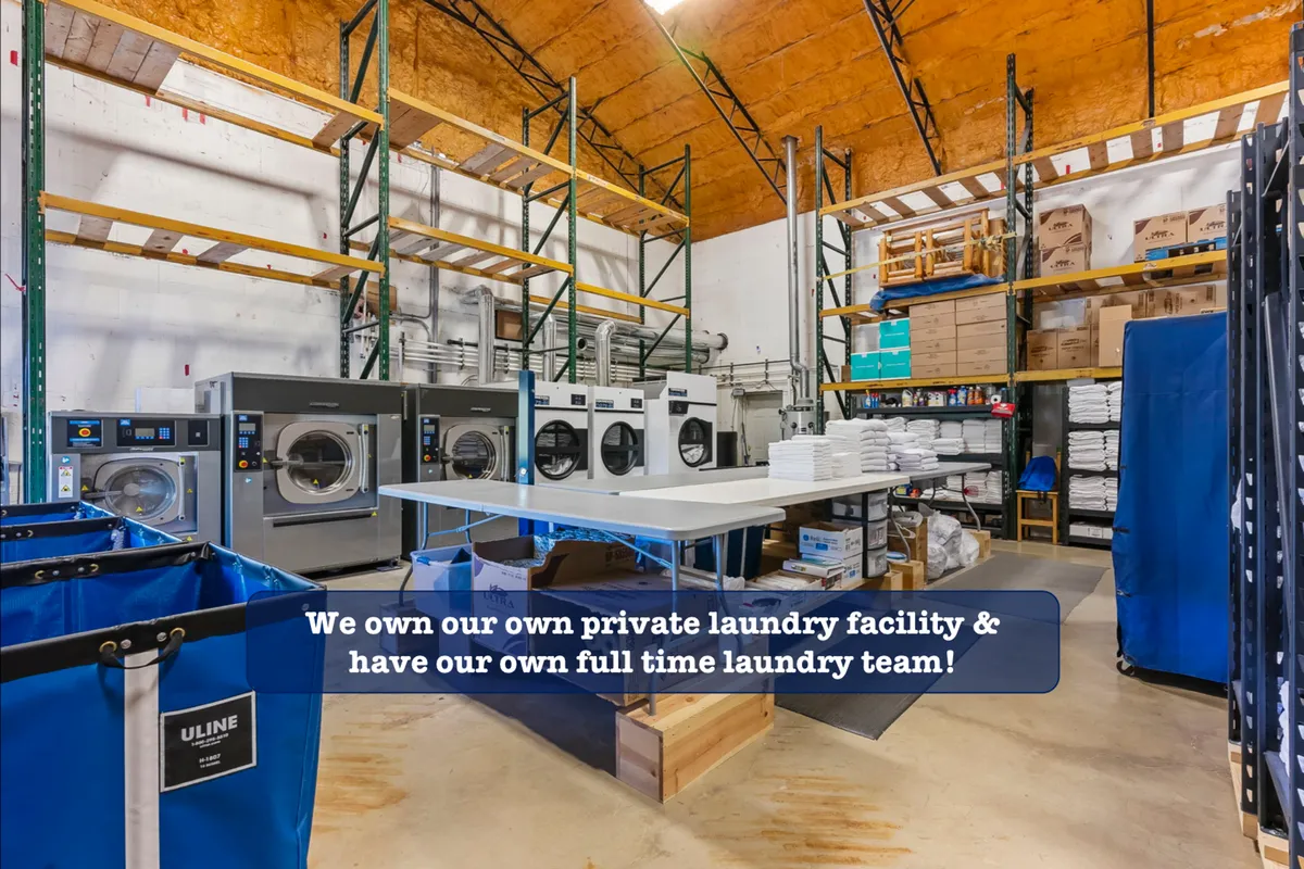 A practical, interior scene showing modern industrial washing machines, storage shelves, laundry carts, and a folding table. The space looks efficient and well-maintained, suggesting convenience for guests handling laundry during their stay.