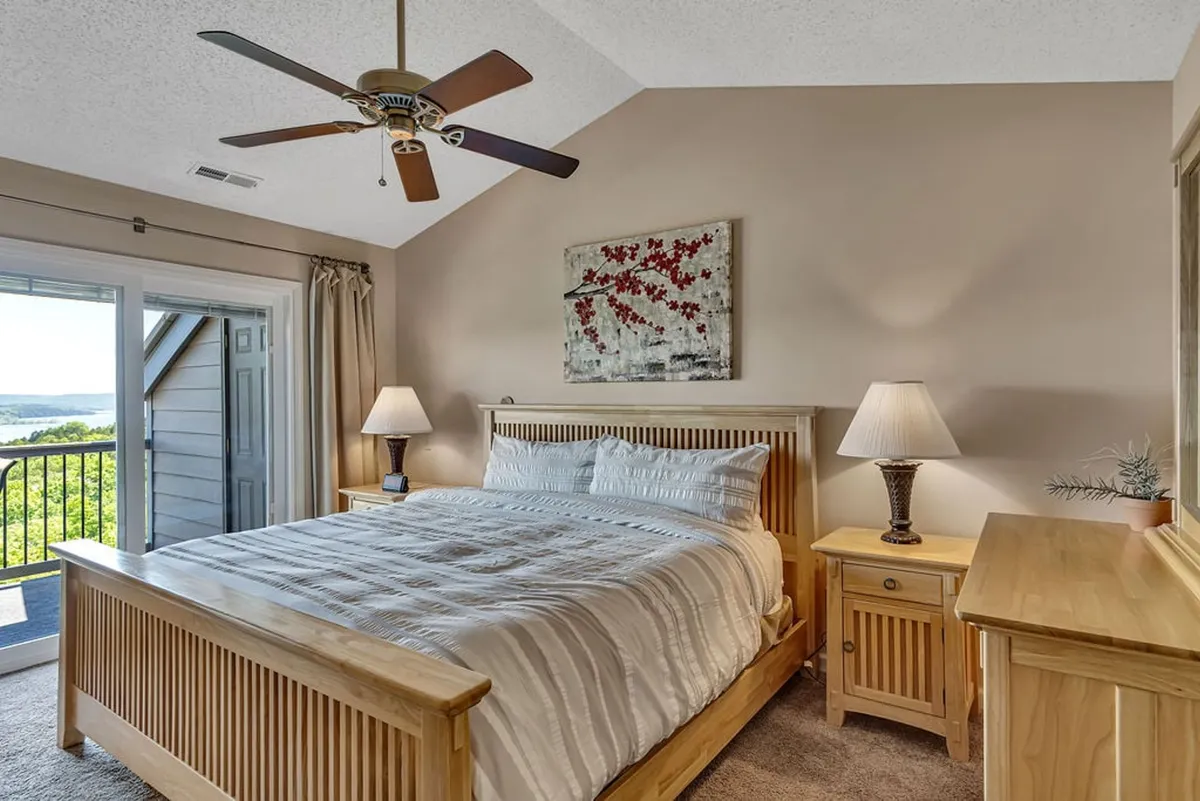 A tranquil master bedroom featuring a king-sized bed with wooden nightstands, tasteful artwork on the wall, and a door leading out to a private balcony. Soft natural light fills the room, creating an inviting and serene atmosphere ideal for rest and relaxation.