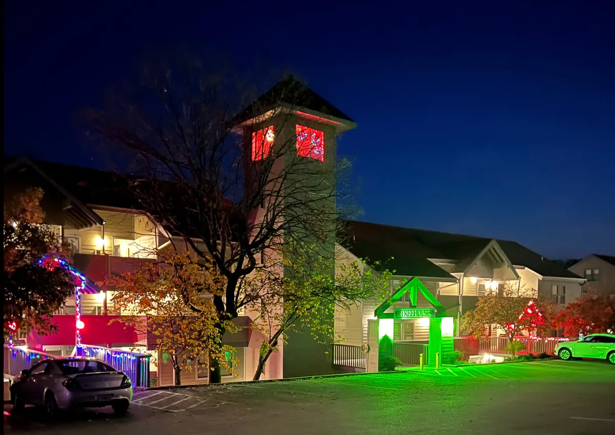 A welcoming exterior view of the condo complex at dusk, adorned with decorative holiday lights and a tall clock tower. The green entrance sign and gentle glow from the lights create a festive, inviting atmosphere, hinting at memorable group arrivals and celebrations.